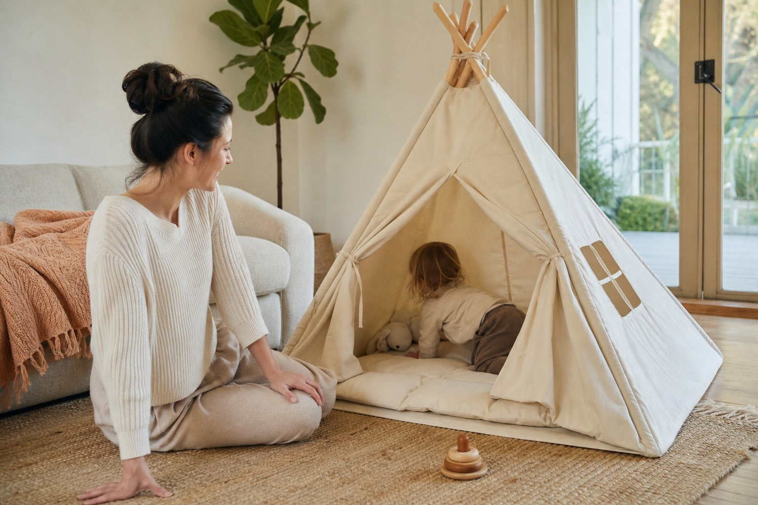 A mother smiling at her toddler crawling into a cream CampCuddle teepee on the living-room floor